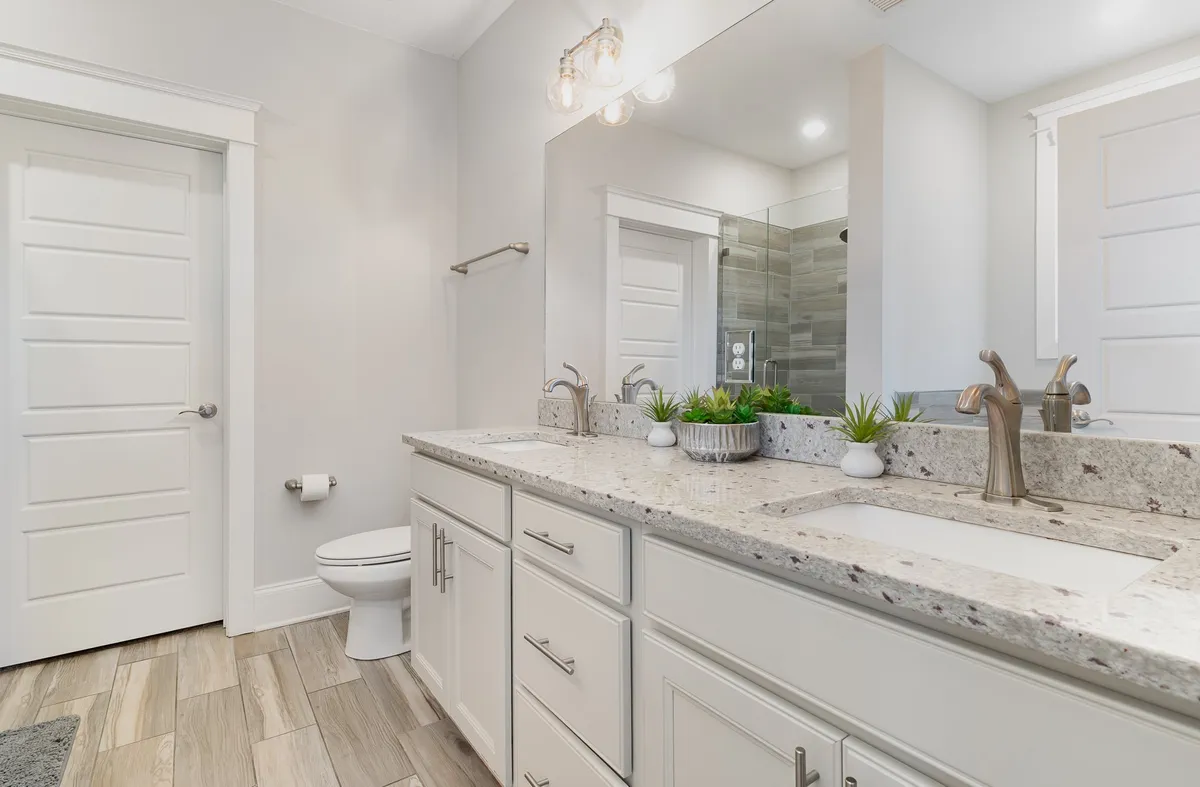 A bright, serene bathroom scene showing a double granite vanity, large mirror, and a spacious shower area. Morning sunlight fills the space, highlighting the elegant fixtures and inviting atmosphere.