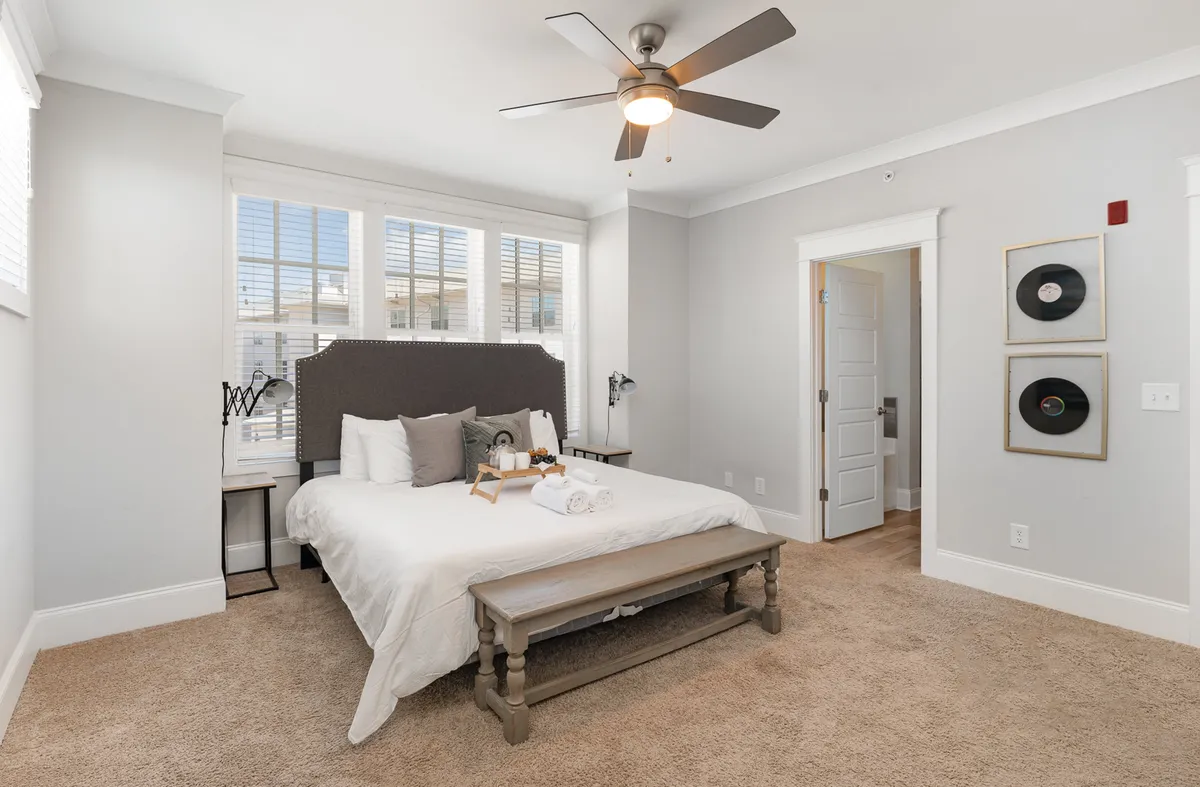 A cozy bedroom featuring a bed adorned with decorative pillows, a wooden bench by the window, and framed records on the wall. Soft light fills the room, emphasizing a comfortable, inviting ambiance.