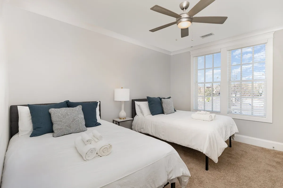 A relaxing bedroom scene with two beds, decorative pillows, a table lamp, and a window with blinds letting in soft daylight. The arrangement suggests ample space for multiple guests, and the dual beds convey versatility for group stays.