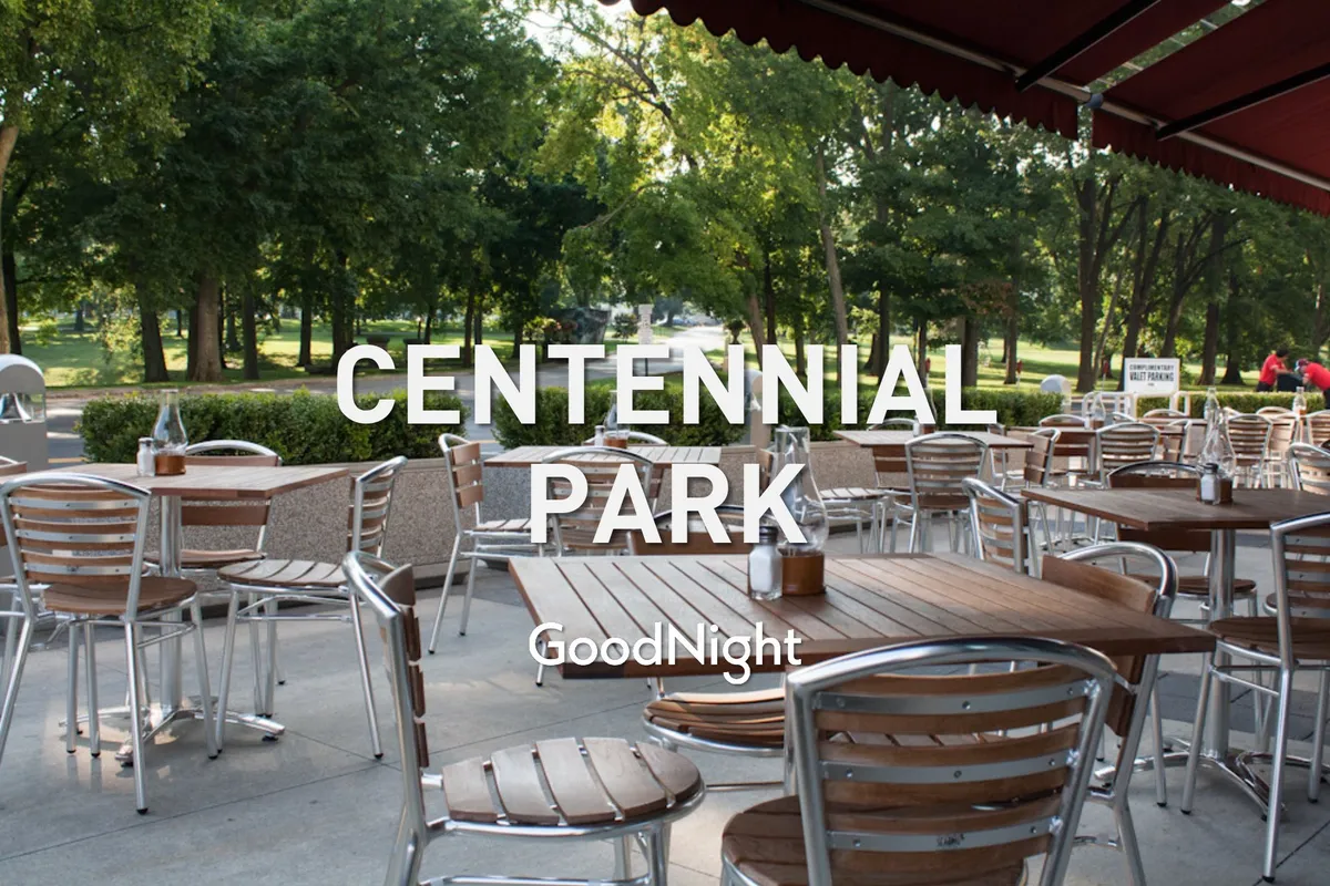 An inviting outdoor dining area set beneath lush green trees, with several tables arranged along a park pathway. The space suggests a relaxed environment ideal for enjoying a meal or gathering with friends after a stroll.