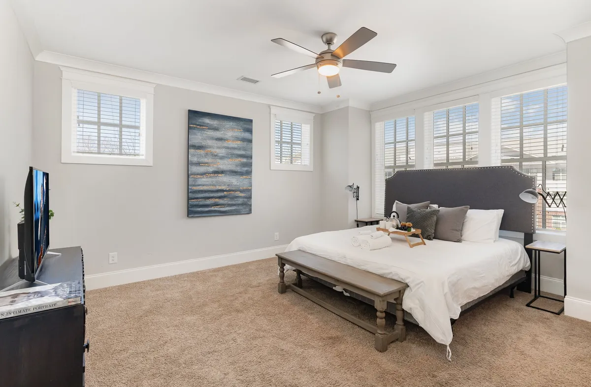 A serene bedroom with a comfortable bed dressed in white linens, tasteful artwork on the wall, a smart TV, and ample natural light streaming in through a window. The mood is peaceful, ideal for relaxation or unwinding with a book or movie.